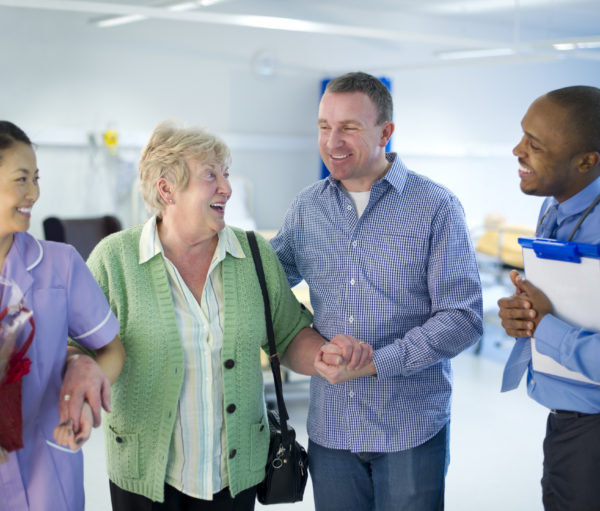 Elderly woman leaving the hospital with son as two hospital staff help and look on