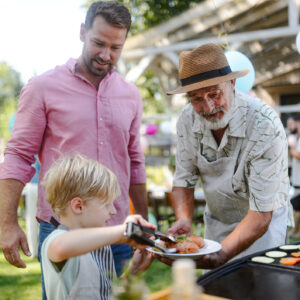 Father, grandfather and son grilling together at a garden bbq party. Three generations of men at summer family garden party.